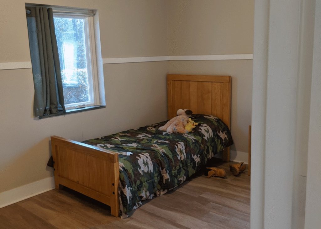 a child's bed in an empty room of an NC PRTF facility. worn stuffed animals on the bed and abandoned untied shoes on the floor. small window is over the bed.