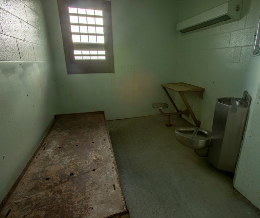 image of a solitary confinement cell - plank to lay on, small window, toilet that is also a sink, and small desk attached to concrete wall