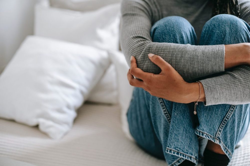 young woman hugging knees sitting on a bed. face is not visible