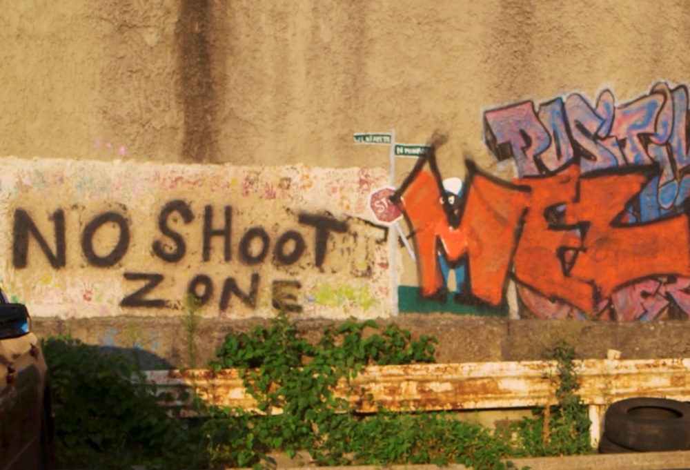 A concrete wall on the side of the road rises up from the ground with graffiti on the right side and the words "No shoot zone" spray painted over a white square to the left of the graffiti