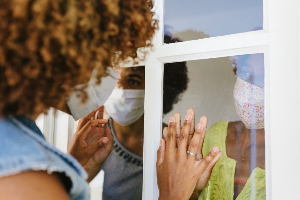 a young woman of color, wearing a face mask , places hands on window and looks in at an older woman of color, wearing a mask who has her hands on the window. Their hands are placed over each other with the window between, symbolizing the isolation and loneliness people with weakened immune systems are facing as they socially distance during the surge of the Delta variant.