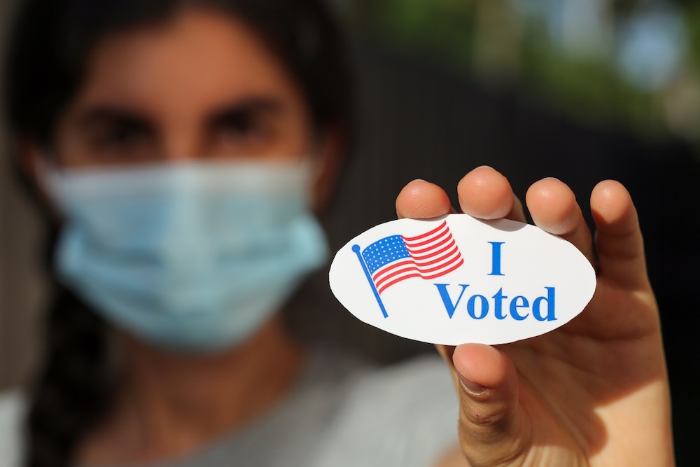 out of focus image of woman with dark brown hair wearing a mask holding up an "I voted" sticker, which is in focus