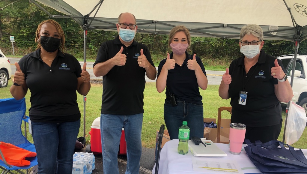 Four staff members of the Piedmont Triad Regional Council who partnered with DRNC on this event stand next to their booth, all wearing black polos and blue jeans, smiling from behind their masks. They all have two thumbs up. From left to right, a Black woman, older white man, a blond-haired woman and a white middle-aged woman.