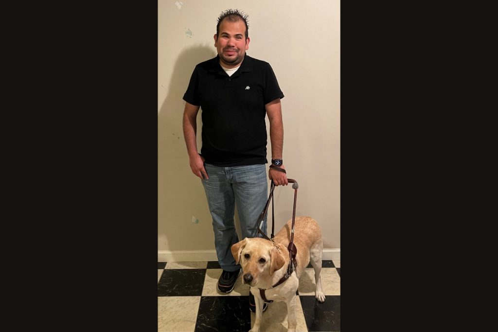 Mr. Oliva, a smiling latino man with black hair, wearing a black polo and Jeans. At his side is his guide dog, Forte, a yellow lab.