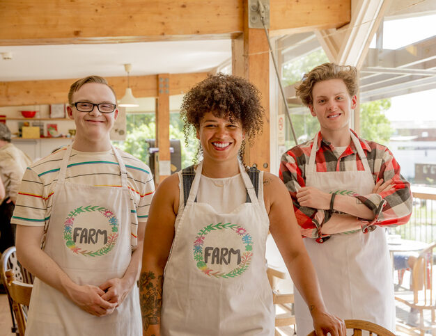 Three people working at the farm cafe pose for a picture. The boss is in the center. She is a person in her 30s with medium brown skin, wearing a "farm" cafe apron and smiling. Two white male employees are on either side of her, also wearing aprons and smiling. One of them has glasses and down syndrome.