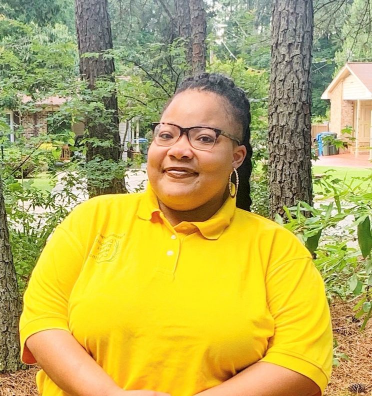 Smiling young black woman wearing glasses and a yellow polo shirt with DRNC logo on it. 