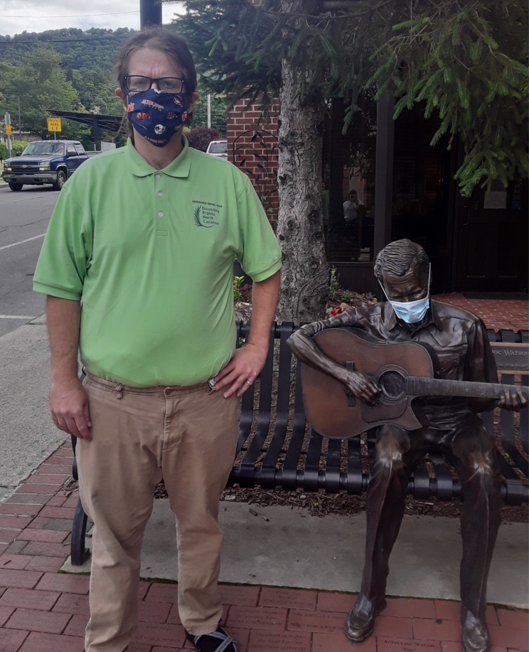 White man wearing green polo with DRNC logo. He has a mask on his face and he is standing next to a bronze guitar-playing statue who is also wearing a mask.