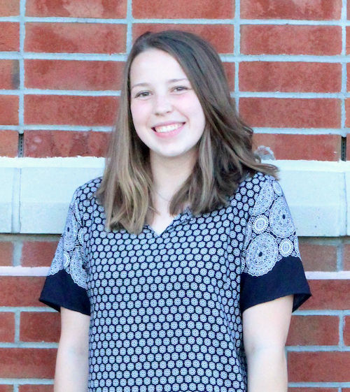 young white woman with light brown hair and brown eyes smiling, wearing blue paisley shirt in front of brick wall