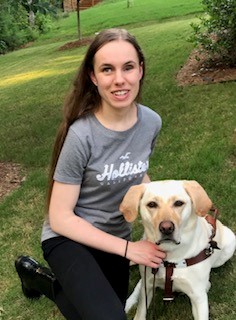 young white woman with long brown hair and brown eyes, smiling while kneeling outside next to her yellow lab guide dog