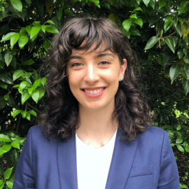 Young white woman with dark brown curly hair wearing blue suit jacket smiling - green leaves of bush behind her