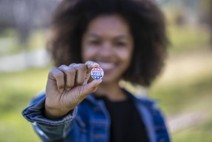 Woman with medium brown skin holds up a blue and red pin that reads "vote." The focus is on her hand, but you can see that she is smiling, has short brown hair, and a jean jacket on. A green background is behind her.