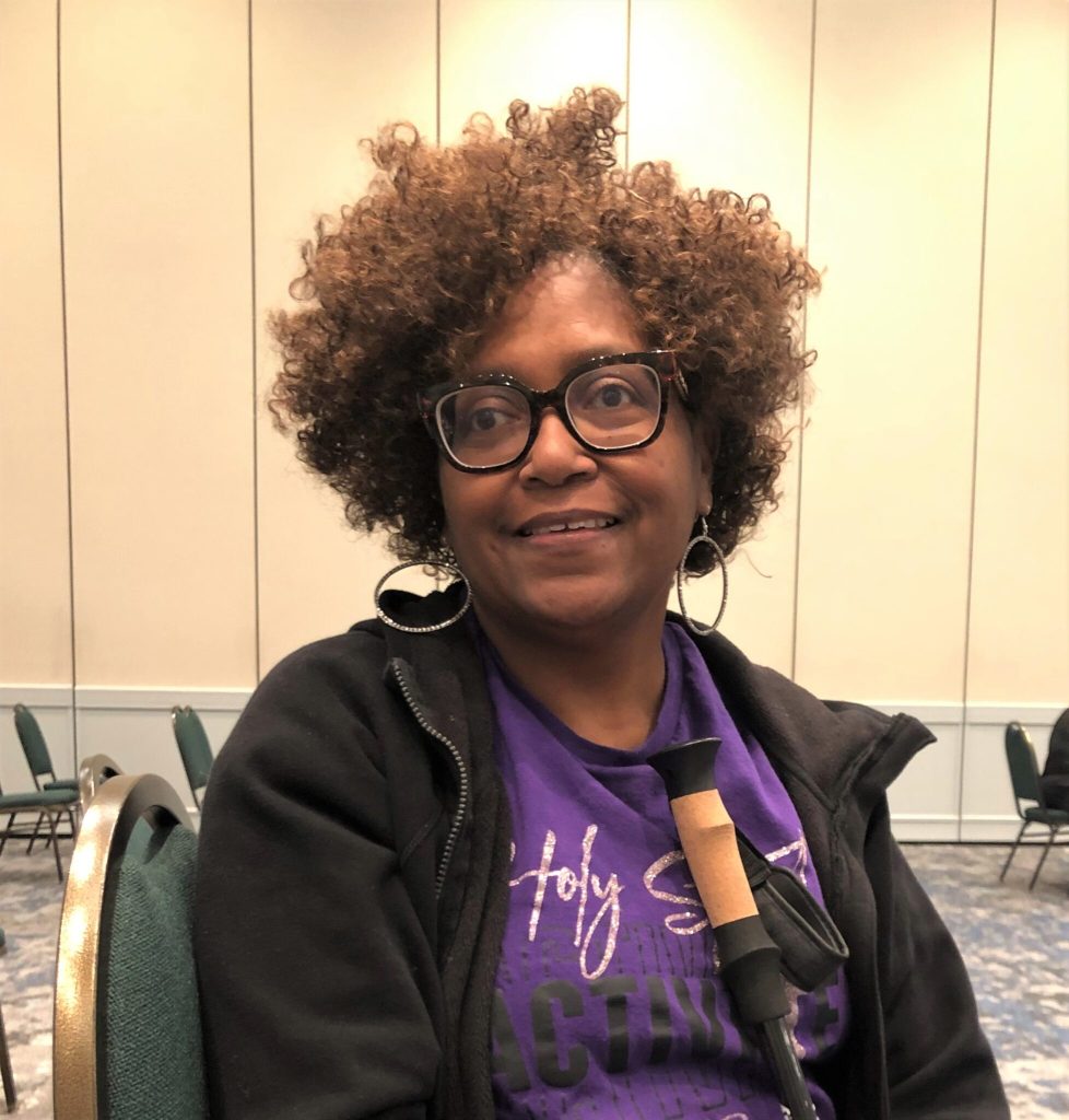 Barbara Lee is smiling at the camera. She is wearing a purple t-shirt and black jacket. She is wearing black framed glasses and holding a cane.
