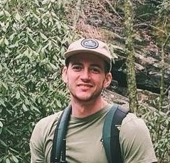 Dane Mullis smiling, wearing a baseball cap and backpack while hiking in the mountains of Western North Carolina