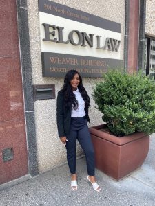 Picture of a young woman with long black hair with a navy suit and a white shirt standing in front of an Elon Law sign. 