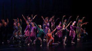 A group of young dancers in bright colored costumes pose on a black stage.