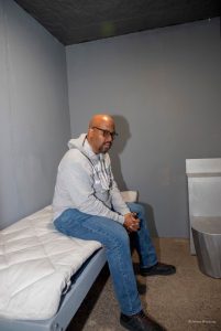 Dr. Craig Waleed is sitting on the bed in the model replica cell.
