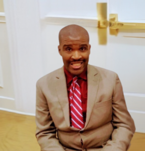 Jermain, a Black man with a close-cropped haircut, looks directly into the camera. He is wearing a tan suit and a red-and-white striped tie