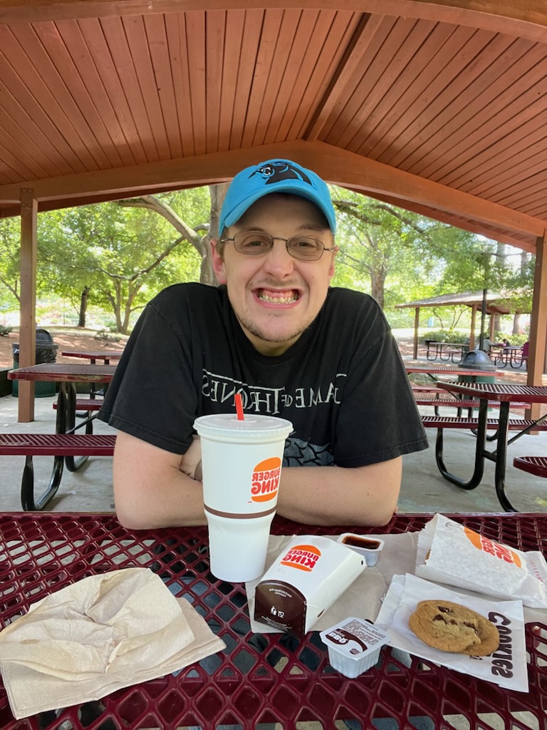 Josh, wearing a bright blue Carolina Panthers baseball cap and a Game of Thrones T-shirt, sits at a picnic shelter with a Burger King lunch in front of him. He is wearing glasses and smiling at the camera.