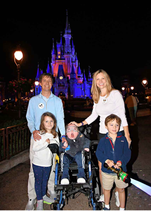 A family of five, including 9-year-old Miles, who is seated in a wheelchair, smiles at the camera. It is nighttime, and Disney World's Cinderella Castle glows in red and blue lights in the background.