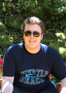 Kellen smiling at the camera on a sunny day, wearing sunglasses and a dark blue Myrtle Beach t-shirt.