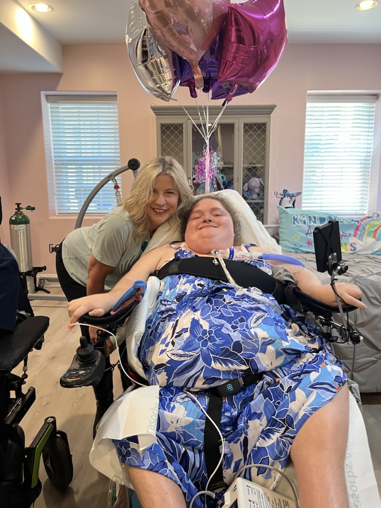 Alexis smiling in the bedroom of her new apartment with DRNC staff member Kirby Morrow. The walls are pink and there are balloons tied to her wheelchair.
