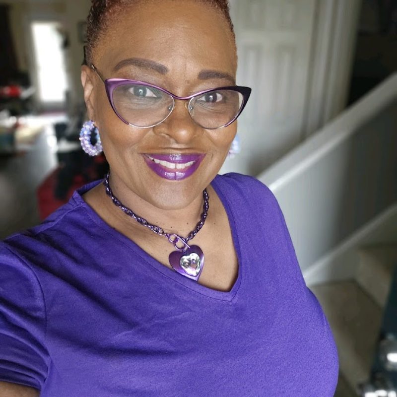 Von Weston wearing a purple shirt and glasses and a purple heartshaped necklace, is smiling at the camera