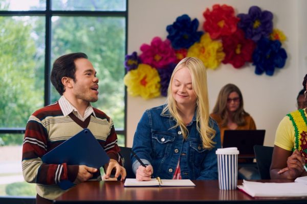 A white professor who has dwarfism stands next to a Caucasian student with Down Syndrome as they have a joyful discussion during class.