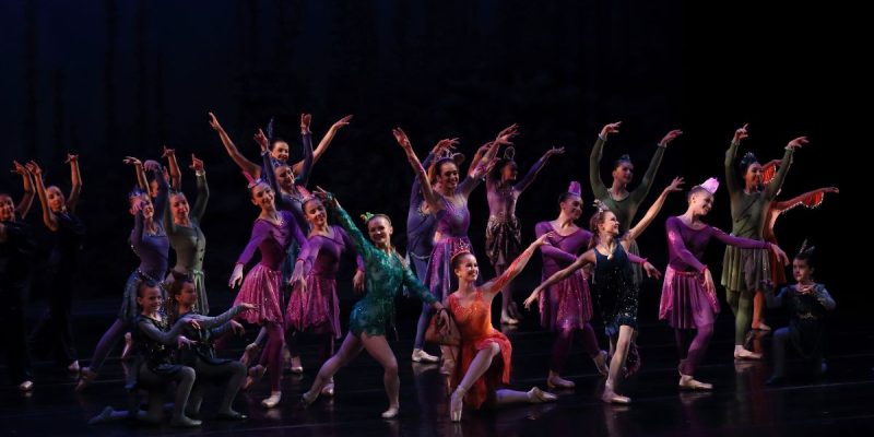 A group of young dancers in bright colored costumes pose on a black stage.