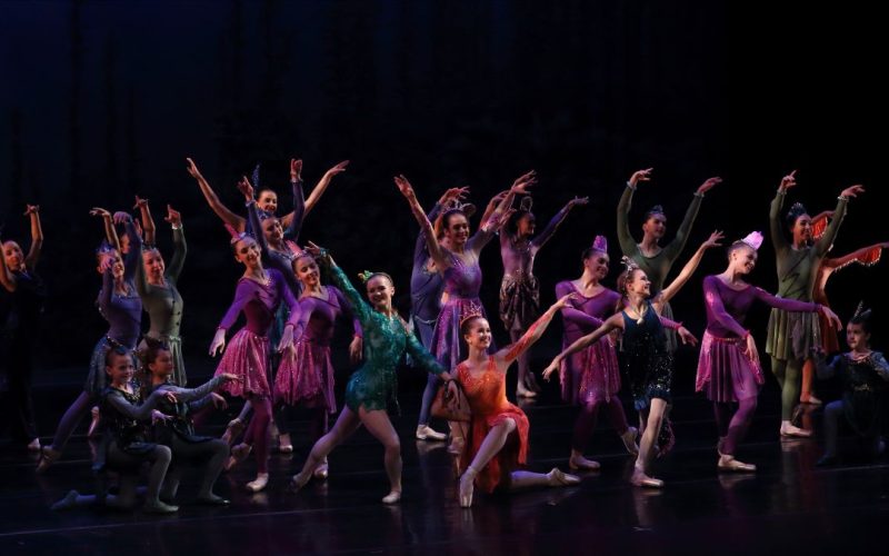 A group of young dancers in bright colored costumes pose on a black stage.