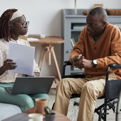Black man in wheelchair meeting with psychologist in ICF-IID facility. The psychologist, who is also black, wears a pink headband, and holds a laptop. She seems to be explaining something to the man.
