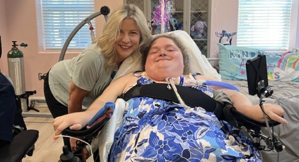 Alexis smiling in the bedroom of her new apartment with DRNC staff member Kirby Morrow. The walls are pink and there are balloons tied to her wheelchair.