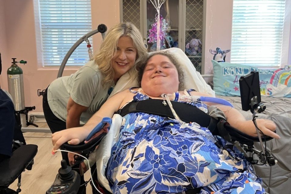 Alexis smiling in the bedroom of her new apartment with DRNC staff member Kirby Morrow. The walls are pink and there are balloons tied to her wheelchair.
