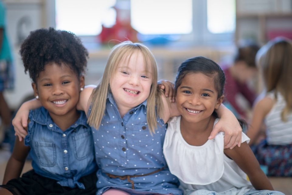 Three multi-ethnic little girls, one with dark brown skin and her curly hair pulled up in a pony, one with blond hair and bangs and light skin, and one with brown skin and braids, are all smiling with their arms across each other's shoulders. They are in a classroom. EPSDT applies to all children with Medicaid.