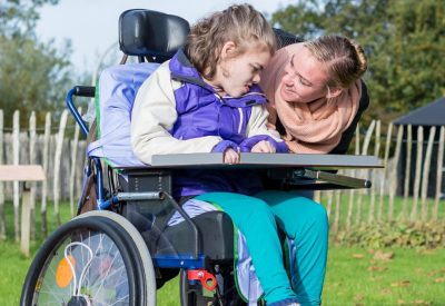 A mom smiles at young daughter in wheelchair outside