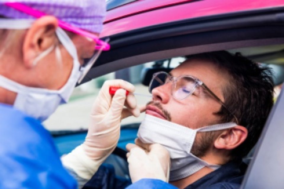 Unvaccinated white man closes eyes as doctor wearing mask and shield tests him for the delta variant through his car window.
