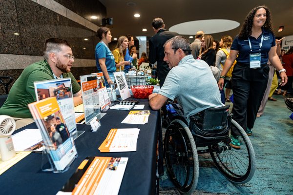 A amn in a wheelchair is talking to a vendor at DRNC's annual conference. Many people are in the background