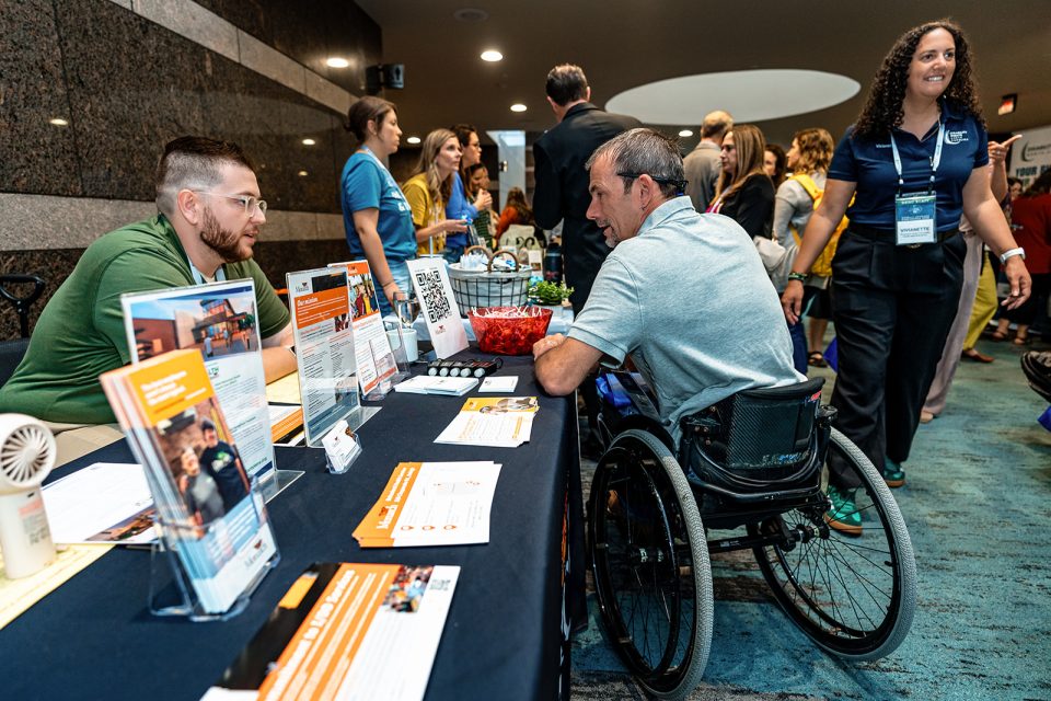 A amn in a wheelchair is talking to a vendor at DRNC's annual conference. Many people are in the background