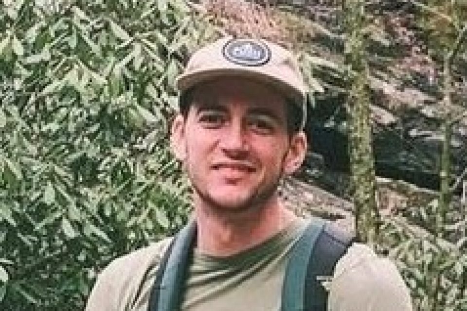 Dane Mullis smiling, wearing a baseball cap and backpack while hiking in the mountains of Western North Carolina