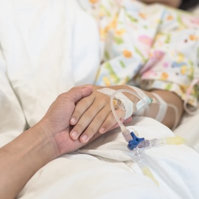 female in hospital bed, arm with an IV showing, a caregiver is holding her hand,