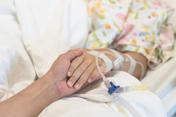 female in hospital bed, arm with an IV showing, a caregiver is holding her hand,
