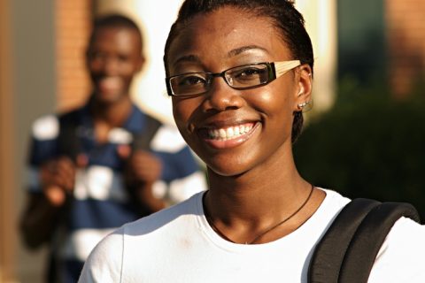 female black high school student wearing glasses and smiling with a back pack slung over her shoulder. Another smiling student appears blurred behind her in the background in front of a school.