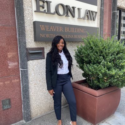 Picture of a young woman with long black hair with a navy suit and a white shirt standing in front of an Elon Law sign.