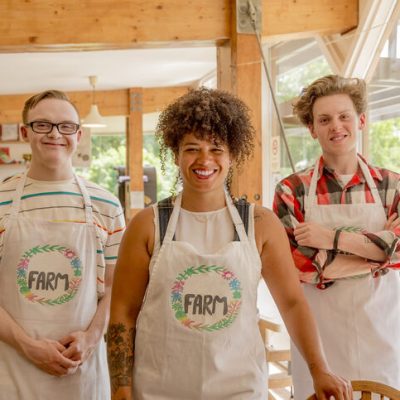 Three people working at the farm cafe pose for a picture. The boss is in the center. She is a person in her 30s with medium brown skin, wearing a "farm" cafe apron and smiling. Two white male employees are on either side of her, also wearing aprons and smiling. One of them has glasses and down syndrome.