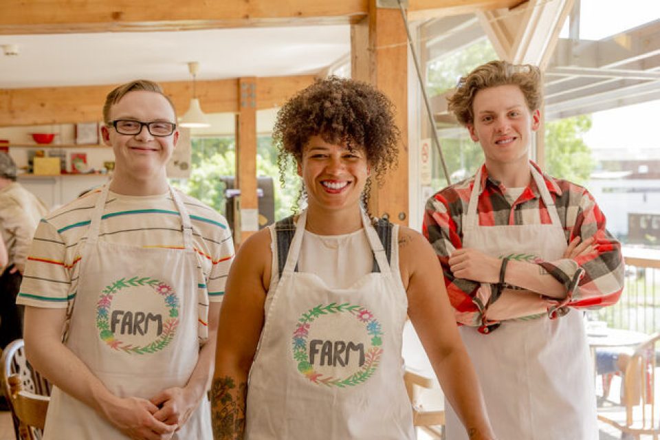 Three people working at the farm cafe pose for a picture. The boss is in the center. She is a person in her 30s with medium brown skin, wearing a 