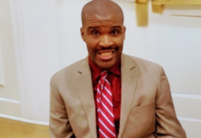 Jermain Jermain, a Black man with a close-cropped haircut, looks directly into the camera. He is wearing a tan suit and a red-and-white striped tie