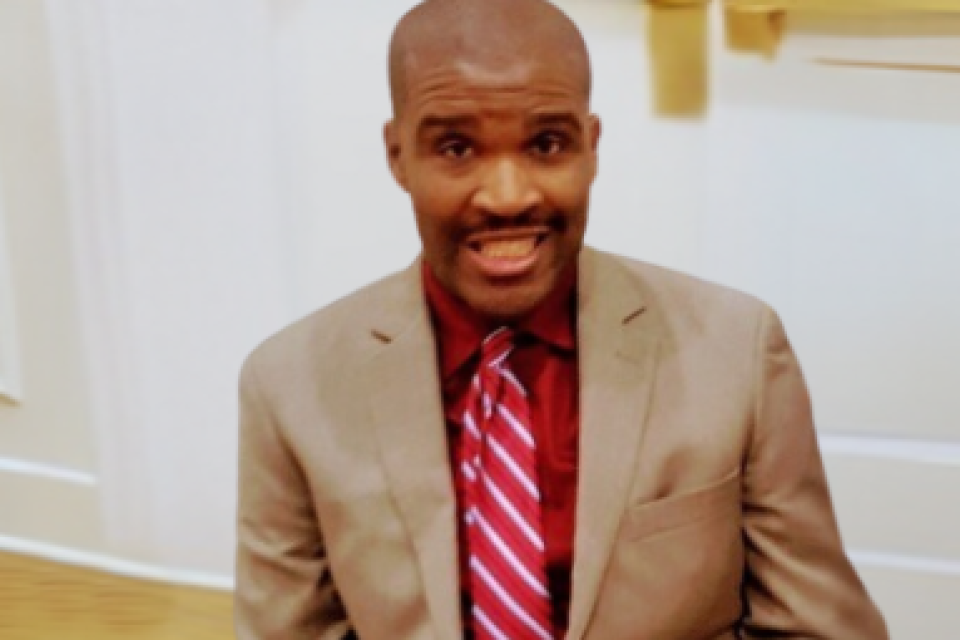 Jermain, a Black man with a close-cropped haircut, looks directly into the camera. He is wearing a tan suit and a red-and-white striped tie