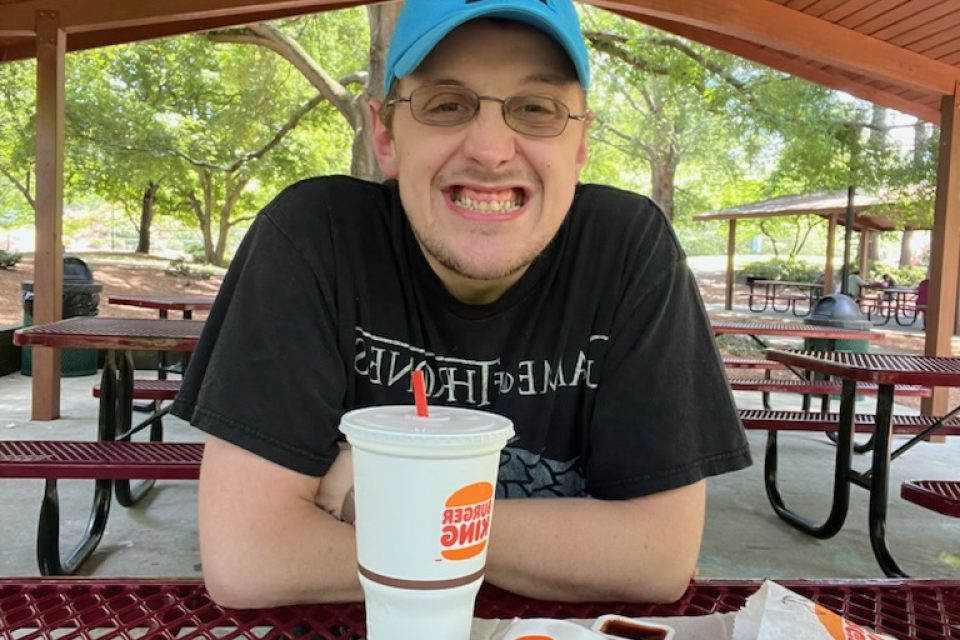Josh, wearing a bright blue Carolina Panthers baseball cap and a Game of Thrones T-shirt, sits at a picnic shelter with a Burger King lunch in front of him. He is wearing glasses and smiling at the camera.