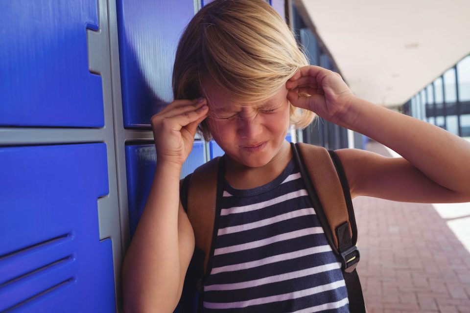 Kid holds his head with his hands in front of blue lockers at school