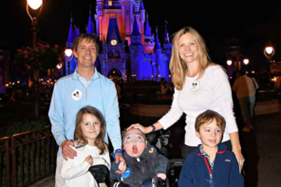 A family of five, including 9-year-old Miles, who is seated in a wheelchair, smiles at the camera. It is nighttime, and Disney World's Cinderella Castle glows in red and blue lights in the background.