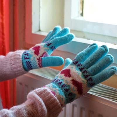 Person with gloves and winter clothes warms hands over a radiator